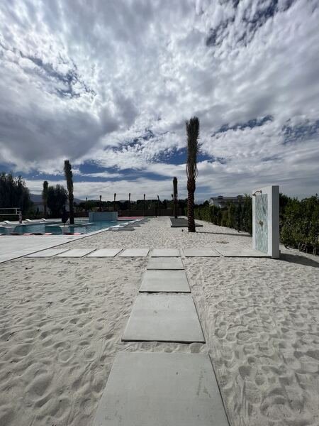 Concrete stepping pads across sand beneath palms toward an outdoor shower