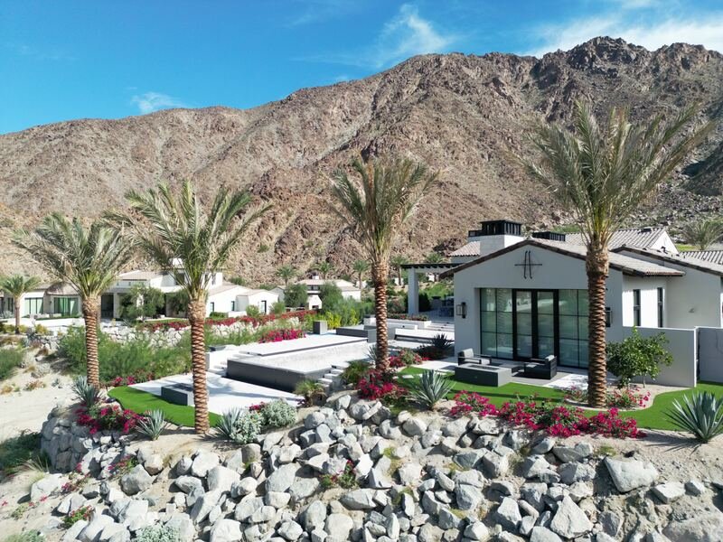 Boulder-lined desert slope with date palms and flowering shrubs below rugged mountains