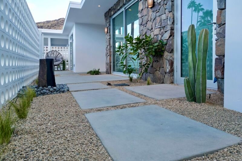 Stone-clad front entry with concrete pads through gravel, a tall totem cactus and a basalt fountain